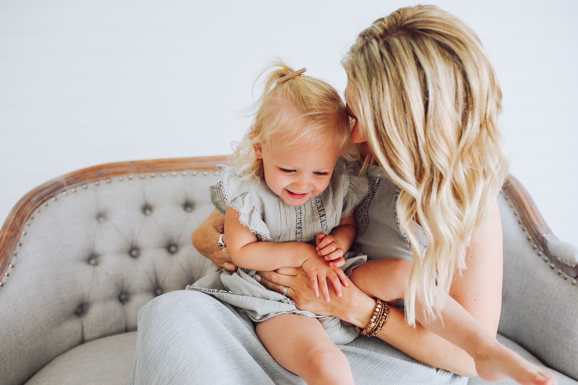 mom with blonde hair tickling smiling blonde hair toddler girl on lap pale blue matching dresses sitting on pale blue buttoned couch